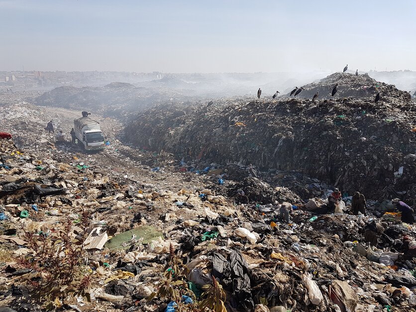 The dump site in Dandora, Kenya. A white truck with people by it sits in the path forged through all the waste. It is a rugged terrain of waste, with trashed piled high into a mountain on the right, atop which birds sit. Strewn about are plastic bags and bottles, but the waste is so great that has created its own terrain.