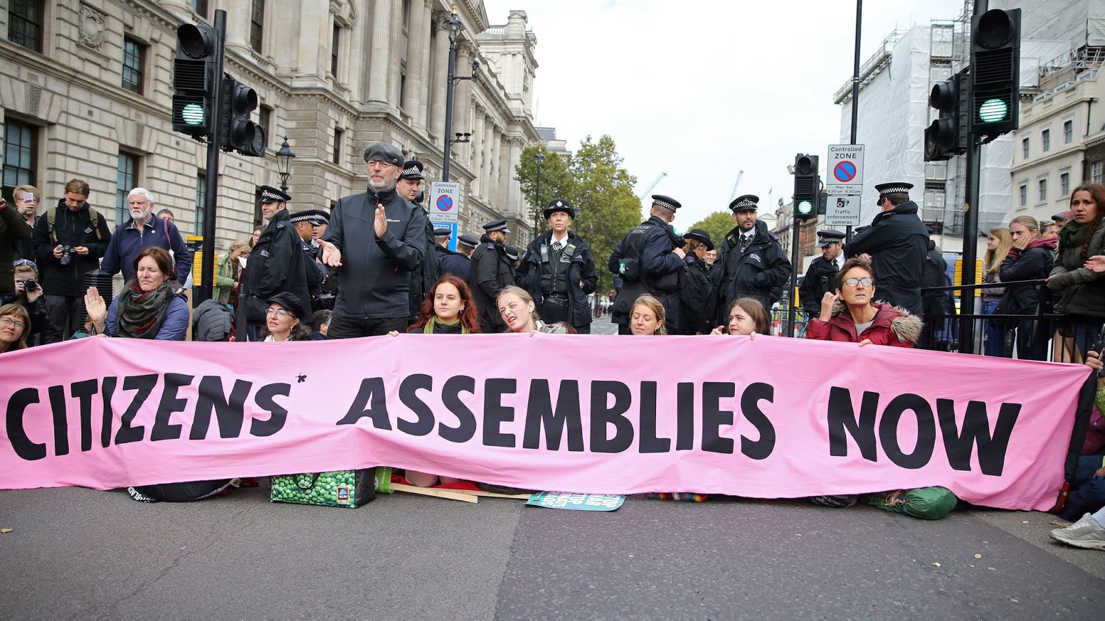 Image of protestors blocking a street with a large banner with the words "citizens' assemblies now" on it.