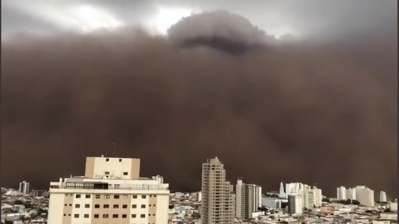 Tall buildings in the foreground are overshadowed by an oncoming wall of thick black smoke around three times their height. 